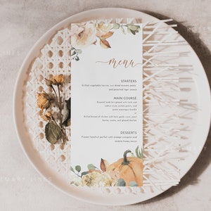 K&ouml;nnte beinhalten: Eine Men&uuml;karte mit floralen und K&uuml;rbis-Illustrationen, pr&auml;sentiert auf einem wei&szlig;en Teller mit einem dekorativen Webmuster. Das Men&uuml; zeigt einen handgeschriebenen Titel "Men&uuml;" und eine Liste von Gerichten.
