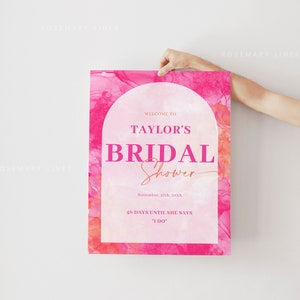 May include: A pink bridal shower sign with the text "Welcome to Taylor's Bridal Shower". The sign includes the date, and the phrase "48 days until she says I do". The background has a watercolor effect.