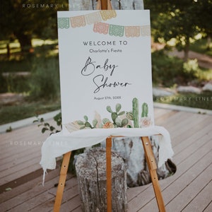Pu&ograve; includere: Un cartello di benvenuto bianco con un design di cactus e fiori. Il cartello recita "Benvenuti alla festa di baby shower di Charlotte August 27th, 20XX".