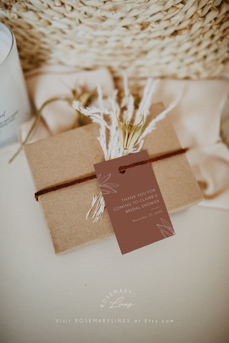 May include: A gift box wrapped in brown paper, tied with a dark brown cord, and adorned with a terracotta-colored tag. The tag reads "THANK YOU FOR COMING TO CLAIRE'S BRIDAL SHOWER November 27, 2020". Dried flowers and foliage add a natural touch.