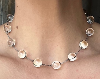 May include: A delicate silver chain necklace with clear glass beads. The necklace is worn by a person with a brown sweater.