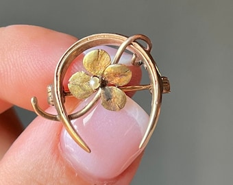 May include: A gold-toned brooch featuring a four-leaf clover design with a small pearl in the center. The brooch is set within a horseshoe-shaped frame. The clover has detailed petals, and the overall design is delicate and vintage.