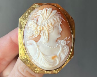 May include: A gold-toned cameo brooch featuring a detailed carving of a woman's profile with a floral headdress. The cameo is set in a gold-toned frame with an intricate design.