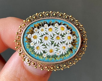 May include: Oval-shaped gold-toned brooch featuring a mosaic of white daisies with yellow centers on a turquoise background. The brooch has a decorative filigree border, showcasing a floral theme. A vintage-inspired accessory.