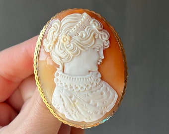 May include: A cameo brooch featuring a carved portrait of a woman with a crown, set in a gold tone oval frame.