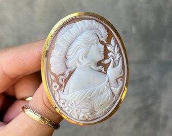 May include: A white cameo brooch with a gold-tone setting. The cameo features a woman's profile with a flower in her hand.