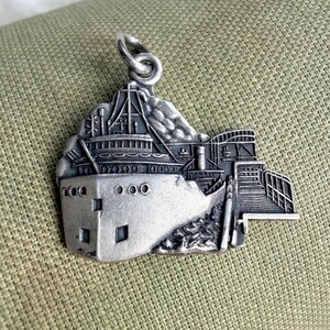 May include: A silver-colored pendant in the shape of a ship with a mountain in the background. The pendant has a detailed design of the ship's structure, including windows and railings. The pendant is attached to a loop for hanging.