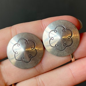 May include: A pair of silver earrings with a flower-shaped design. The earrings are round and have a raised center with five small circles around it.