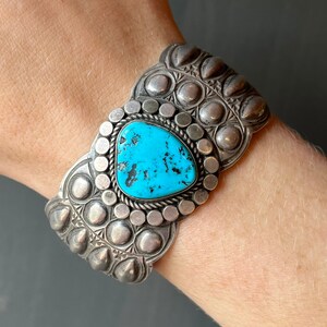 May include: A silver cuff bracelet with a turquoise stone set in the center. The bracelet is decorated with a repeating pattern of small circles and raised bumps.