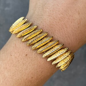 May include: A gold-toned bracelet with an articulated design. The bracelet features elongated, leaf-shaped links with floral and etched patterns. The bracelet is worn on a wrist, showcasing its intricate details and warm metallic color.