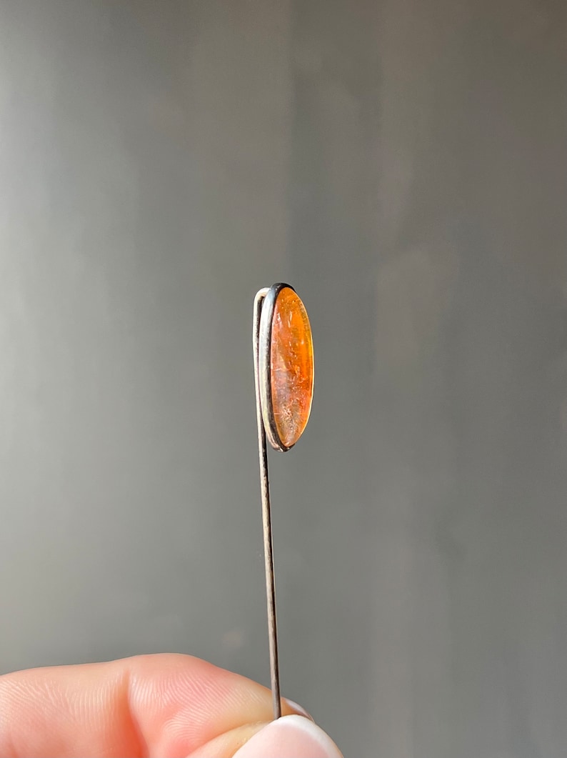 May include: A silver tie tack with an oval-shaped orange stone.