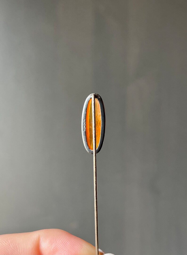 May include: A silver tie tack with an oval-shaped amber stone. The back of the tie tack is marked "Sterling".