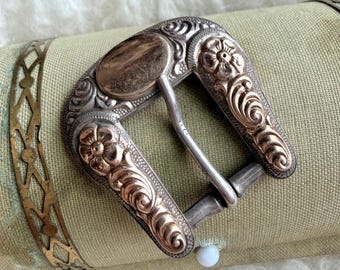 May include: A belt buckle featuring a two-tone design with silver-colored metal and rose gold-colored floral engravings. The buckle has a rounded rectangular shape with a central prong and a smooth, unadorned oval section at the top.