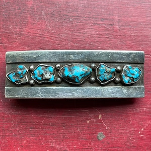 May include: A silver belt buckle with a row of turquoise stones set in silver. The stones are a variety of shapes and sizes, and they are all a vibrant blue color.