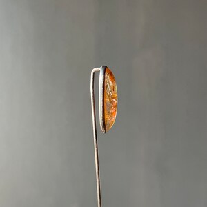 May include: A close-up of a small, oval-shaped, orange and brown gemstone set in a silver setting. The gemstone is attached to a thin silver wire.