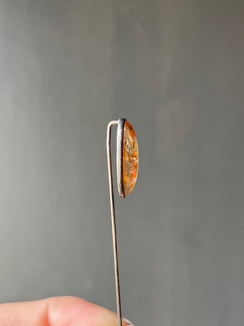 May include: A close-up of a small, oval-shaped, orange and brown gemstone set in a silver setting. The gemstone is attached to a thin silver wire.