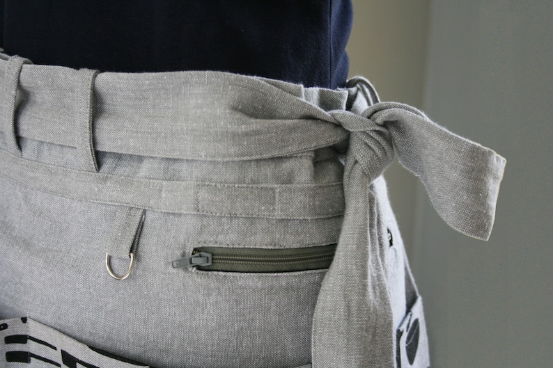 May include: Close-up of a light gray apron with a tied waist. The apron features a zippered pocket, a metal ring, and a decorative pattern on the lower portion. The fabric appears to be linen or a similar material.