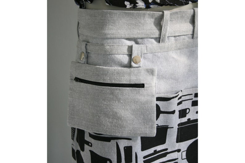 May include: A gray apron with a black zipper pocket and a black and white kitchen utensil pattern on the lower portion. The apron has a gray waistband with silver snap closures.