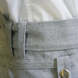 May include: Close-up of a gray fabric waistband with a button and belt loops. The waistband is made of a textured material and is paired with a white shirt.