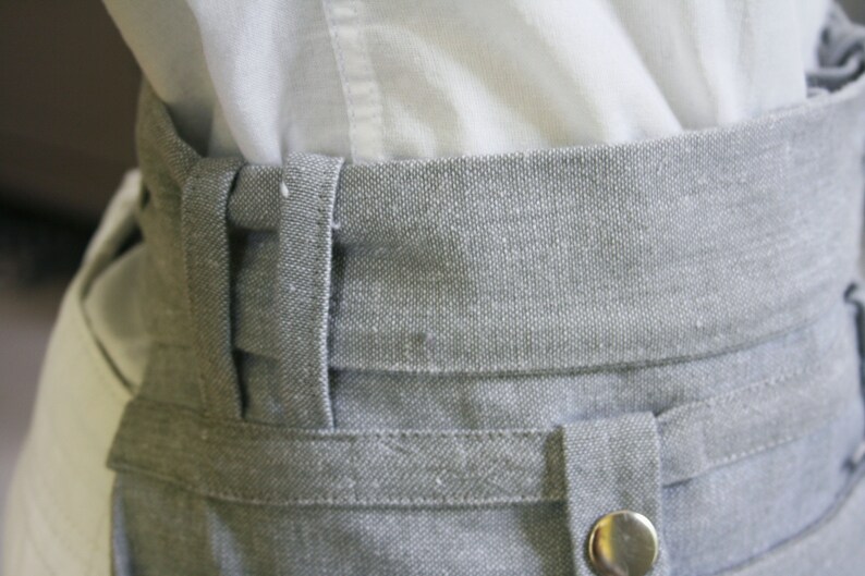 May include: Close-up of a gray fabric waistband with a button and belt loops. The waistband is made of a textured material and is paired with a white shirt.