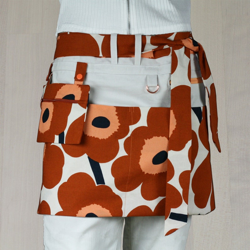 May include: A waist apron with a floral pattern in shades of brown, peach, and black on a white background. The apron has a light gray pocket and a small matching pouch. The apron ties at the waist.