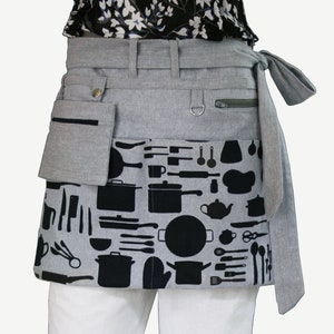 May include: A gray waist apron with black kitchen utensil and cookware silhouettes. Features include a front pocket, a zippered pocket, and a D-ring. The apron ties at the waist and is designed for use in the kitchen.