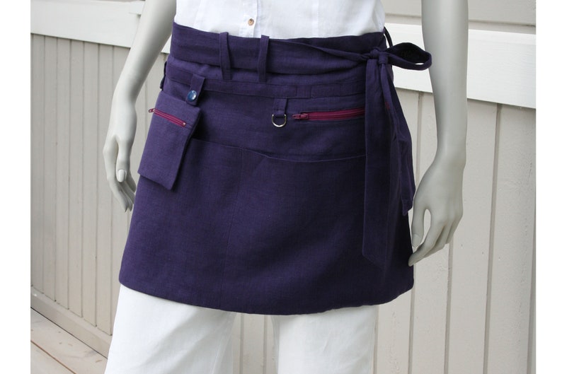 May include: A dark purple apron with multiple pockets and a wrap-around tie. The apron features a small pocket with a button closure, a zippered pocket with a red zipper, and a metal ring detail. The apron is made of a textured fabric.