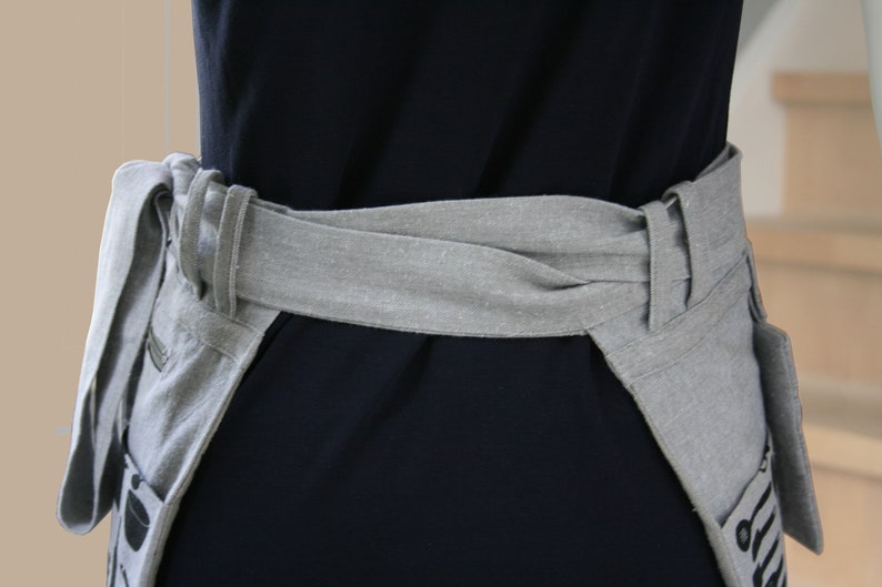 May include: A gray apron with a wrap-around waist tie, featuring a pocket with a black and white design. The apron is made of a light-colored fabric and is worn over a dark blue garment.