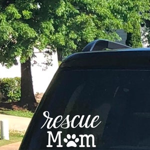 May include: A white vinyl decal on a car window. The decal reads "rescue Mom" in a cursive font, with the word "Mom" below in a block font, and a paw print replacing the "o". The background shows green trees.