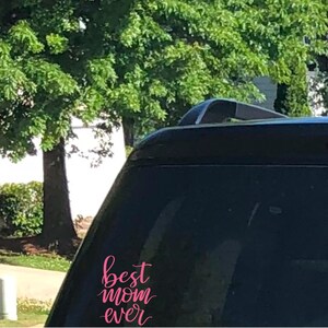May include: A black vehicle with the words "best mom ever" in pink script on the rear window. The background features green trees and a bright sunny day.