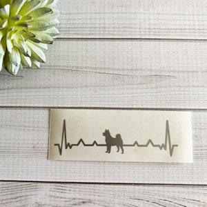 May include: A gray decal featuring a dog silhouette within a heartbeat line. The design is on a rectangular sticker. The background is a light wood grain pattern, with a green succulent in the upper left corner.