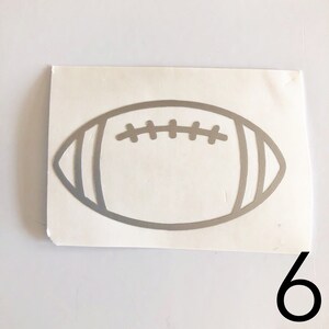 May include: A silver-colored football-shaped sticker with white backing. The sticker features the outline of a football with detailed stitching and stripes. The number 6 is in the bottom right corner.