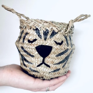 Puede incluir: Una cesta tejida con un diseño de cara de tigre. La cesta está hecha de paja de color natural y presenta rayas negras y una nariz negra. La cesta tiene dos asas pequeñas y está sostenida en la mano de una persona.