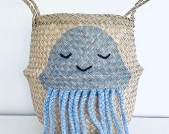 May include: Woven seagrass basket with a hand-painted blue jellyfish design. The jellyfish has a smiling face and blue yarn tentacles. The basket has two woven handles.
