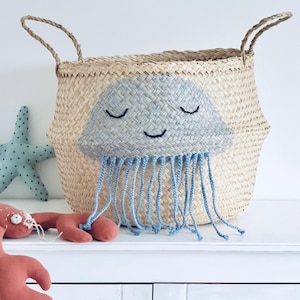 Puede incluir: Cesta de almacenamiento de hierba marina tejida con un diseño de medusa sonriente pintado a mano en azul claro y gris. La cesta tiene dos asas y flecos azules. Se ven otros juguetes de peluche.