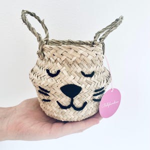 May include: Woven basket with a cat face design. The basket is made of natural-coloured straw and features a black nose, whiskers, and closed eyes. It has two woven handles and a pink tag that says "Bellybambino".