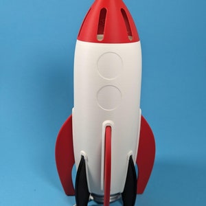 May include: A red, white, and black rocket-shaped lamp with a cone-shaped top and fins. The lamp has a white body with two circular cutouts and a red top with ventilation slots. A white power cord extends from the base.