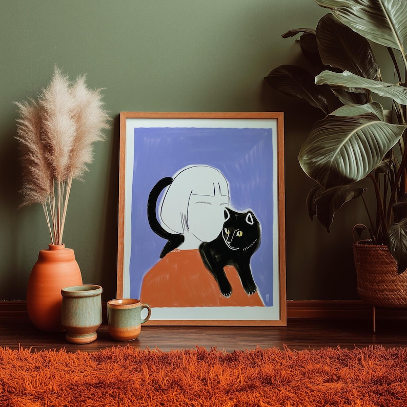 May include: A framed art print featuring a stylized illustration of a person with white hair and a black cat on their shoulder. The artwork has a blue background and is in a wooden frame. The room has a terracotta vase with pampas grass, two ceramic mugs, and an orange rug.