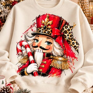 May include: A cream-colored crewneck sweatshirt featuring a vibrant graphic print of a nutcracker. The nutcracker wears a red uniform with gold fringe epaulets, a tall red hat with a leopard print bow, and holds a red and white candy cane.