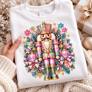 May include: A white sweatshirt featuring a central graphic of a nutcracker soldier in iridescent pink, blue, and gold tones. The nutcracker is surrounded by pine branches, pink and blue Christmas ornaments, bows, and snowflakes.