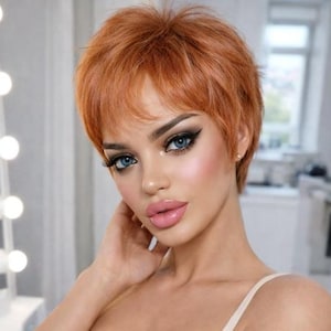 May include: A woman with short, red hair and blue eyes, wearing a beige tank top. The image includes a logo with the text "ELESSA" and a crown design. The background is a blurred interior space with a vanity mirror.