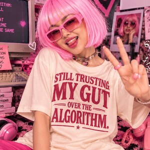 Puede incluir: Una camiseta de manga corta color crema con el texto en rojo "STILL TRUSTING MY GUT OVER THE ALGORITHM." La prenda se combina con pantalones de color rosa brillante, gafas de sol rectangulares tintadas de rosa, un collar de pinchos plateado y un anillo rosa.