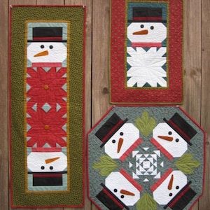 Könnte beinhalten: Drei handgefertigte Schneemann-Quilts in verschiedenen Formen. Einer ist rechteckig mit einem grünen Rand und einem Schneemann-Design. Ein weiterer ist rechteckig mit einem roten Rand. Der dritte ist ein Achteck mit vier Schneemännern.