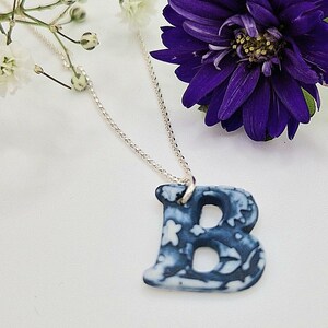 Puede incluir: Un collar de cadena de plata con un colgante de cerámica en forma de letra B con estampado azul y blanco.