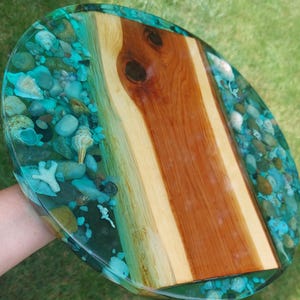 May include: A round serving board featuring a central piece of natural wood with a prominent knot, flanked by two sections of clear turquoise resin embedded with various smooth river stones, small white seashells, and coral-like fragments.