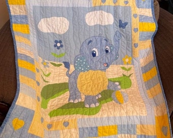 May include: A blue and yellow baby quilt with a cartoon elephant, clouds, and flowers. The quilt has a striped border and a heart pattern.
