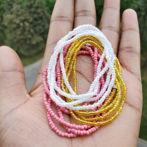 Pu&ograve; includere: Una collezione di tre fili di perline sovrapposti con piccole perline di vetro rotonde, bianche opache, gialle traslucide e rosa a tinta unita. Ogni filo &egrave; fissato con una piccola chiusura in metallo color argento.
