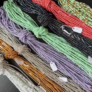 Pu&ograve; includere: Molteplici fili di piccole perline di vetro in vari colori, tra cui verde chiaro, corallo, blu scuro, lavanda, argento, oro e bianco. Ogni fascio &egrave; legato con un nodo e presenta una piccola etichetta bianca con la scritta 28 inches.