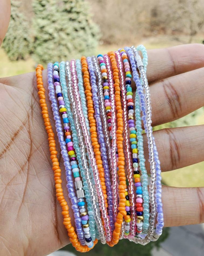 Pu&ograve; includere: Una collezione di diverse collane o braccialetti sottili composti da piccole perline di vetro colorate nei toni dell'arancione, lavanda, azzurro, rosa, giallo e bianco, con chiusure a barilotto in metallo argentato che fissano i fili.