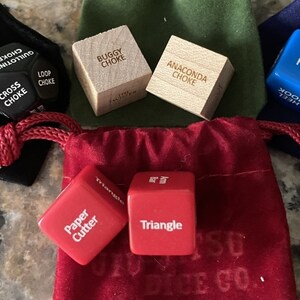 May include: A set of six dice with various phrases printed on each side. The dice are in different colors, including black, blue, red, and wood. The dice are in a red velvet bag with the words "SU DICE CO." printed on it.